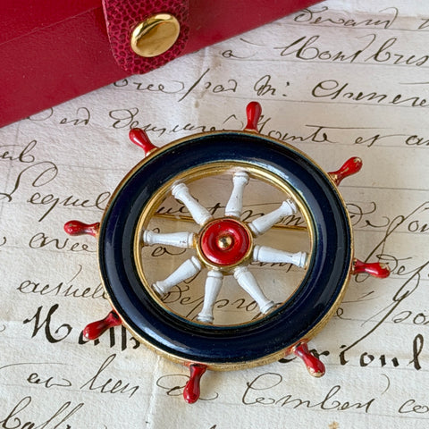 Vintage gold‑tone ship‑wheel brooch with navy, red, and white enamel, photographed on antique French script background.