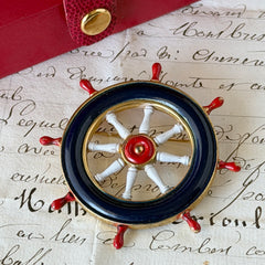 Vintage gold‑tone ship‑wheel brooch with navy, red, and white enamel, photographed on antique French script background.