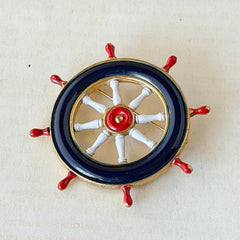 Vintage gold‑tone ship‑wheel brooch with navy, red, and white enamel, photographed on antique French script background.