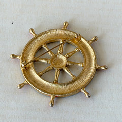 Vintage gold‑tone ship‑wheel brooch with navy, red, and white enamel, photographed on antique French script background.