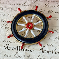 Vintage gold‑tone ship‑wheel brooch with navy, red, and white enamel, photographed on antique French script background.