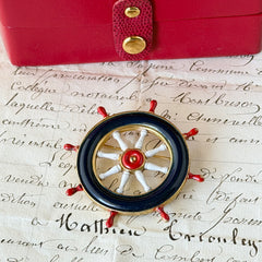Vintage gold‑tone ship‑wheel brooch with navy, red, and white enamel, photographed on antique French script background.