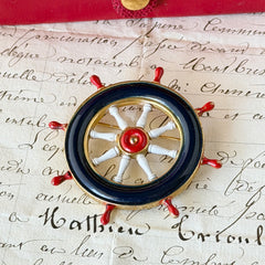 Vintage gold‑tone ship‑wheel brooch with navy, red, and white enamel, photographed on antique French script background.