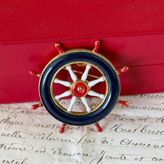 Vintage gold‑tone ship‑wheel brooch with navy, red, and white enamel, photographed on antique French script background.