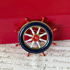 Vintage gold‑tone ship‑wheel brooch with navy, red, and white enamel, photographed on antique French script background.