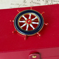 Vintage gold‑tone ship‑wheel brooch with navy, red, and white enamel, photographed on antique French script background.