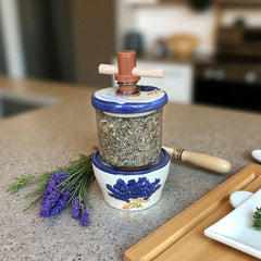 Provence ceramic herb mill with hand-painted blue lavender sprigs and green leaves on a cream base, featuring a glass chamber filled with dried herbs and a wooden crossbar handle for grinding.