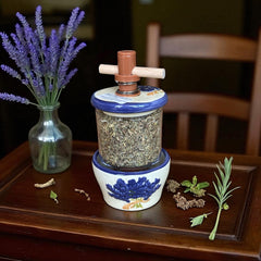 Provence ceramic herb mill with hand-painted blue lavender sprigs and green leaves on a cream base, featuring a glass chamber filled with dried herbs and a wooden crossbar handle for grinding.