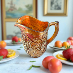 Vintage marigold Carnival Glass pitcher with iridescent grape and leaf pattern, scalloped rim, and curved handle; warm amber tones ideal for fall decor or collector display.
