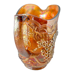 Vintage marigold Carnival Glass pitcher with iridescent grape and leaf pattern, scalloped rim, and curved handle; warm amber tones ideal for fall decor or collector display.