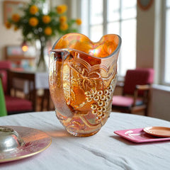 Vintage marigold Carnival Glass pitcher with iridescent grape and leaf pattern, scalloped rim, and curved handle; warm amber tones ideal for fall decor or collector display.