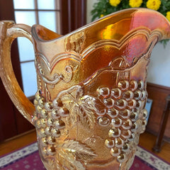Vintage marigold Carnival Glass pitcher with iridescent grape and leaf pattern, scalloped rim, and curved handle; warm amber tones ideal for fall decor or collector display.