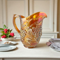 Vintage marigold Carnival Glass pitcher with iridescent grape and leaf pattern, likely Imperial Glass, featuring warm amber, rose, and copper tones; approx. 8¼" tall, in very good condition with minor chips noted on base and rim, ideal for fall decor or vintage display.