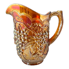 Vintage marigold Carnival Glass pitcher with iridescent grape and leaf pattern, scalloped rim, and curved handle; warm amber tones ideal for fall decor or collector display.