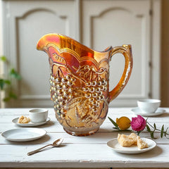 Vintage marigold Carnival Glass pitcher with iridescent grape and leaf pattern, scalloped rim, and curved handle; warm amber tones ideal for fall decor or collector display.