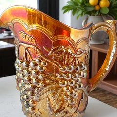 Vintage marigold Carnival Glass pitcher with iridescent grape and leaf pattern, scalloped rim, and curved handle; warm amber tones ideal for fall decor or collector display.