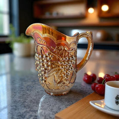 Vintage marigold Carnival Glass pitcher with iridescent grape and leaf pattern, scalloped rim, and curved handle; warm amber tones ideal for fall decor or collector display.