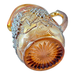 Vintage marigold Carnival Glass pitcher with iridescent grape and leaf pattern, scalloped rim, and curved handle; warm amber tones ideal for fall decor or collector display.