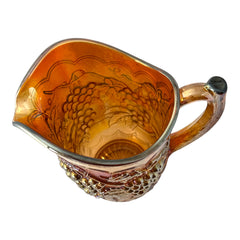 Vintage marigold Carnival Glass pitcher with iridescent grape and leaf pattern, scalloped rim, and curved handle; warm amber tones ideal for fall decor or collector display.