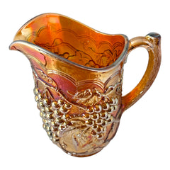 Vintage marigold Carnival Glass pitcher with iridescent grape and leaf pattern, likely Imperial Glass, featuring warm amber, rose, and copper tones; approx. 8¼" tall, in very good condition with minor chips noted on base and rim, ideal for fall decor or vintage display.