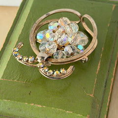 Vintage gold tone brooch featuring a cluster of iridescent aurora borealis crystals framed by curved scrolls and rhinestone accents, shown against various backgrounds.