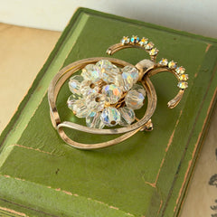 Vintage gold tone brooch featuring a cluster of iridescent aurora borealis crystals framed by curved scrolls and rhinestone accents, shown against various backgrounds.