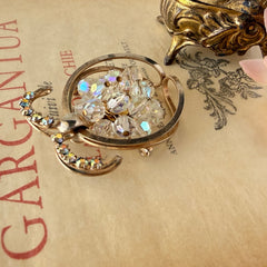 Vintage gold tone brooch featuring a cluster of iridescent aurora borealis crystals framed by curved scrolls and rhinestone accents, shown against various backgrounds.