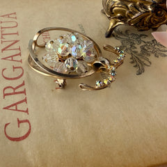 Vintage gold tone brooch featuring a cluster of iridescent aurora borealis crystals framed by curved scrolls and rhinestone accents, shown against various backgrounds.