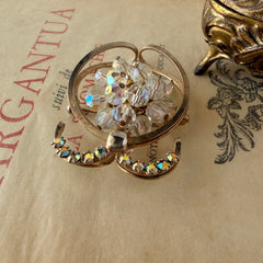 Vintage gold tone brooch featuring a cluster of iridescent aurora borealis crystals framed by curved scrolls and rhinestone accents, shown against various backgrounds.