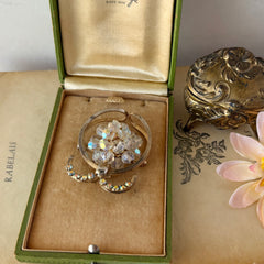 Vintage gold tone brooch featuring a cluster of iridescent aurora borealis crystals framed by curved scrolls and rhinestone accents, shown against various backgrounds.