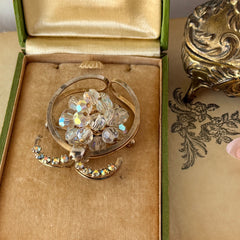 Vintage gold tone brooch featuring a cluster of iridescent aurora borealis crystals framed by curved scrolls and rhinestone accents, shown against various backgrounds.