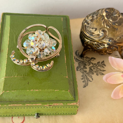 Vintage gold tone brooch featuring a cluster of iridescent aurora borealis crystals framed by curved scrolls and rhinestone accents, shown against various backgrounds.