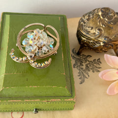 Vintage gold tone brooch featuring a cluster of iridescent aurora borealis crystals framed by curved scrolls and rhinestone accents, shown against various backgrounds.