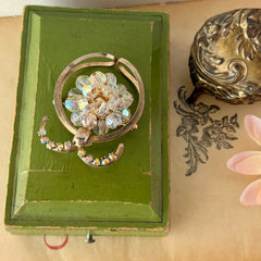 Vintage gold tone brooch featuring a cluster of iridescent aurora borealis crystals framed by curved scrolls and rhinestone accents, shown against various backgrounds.