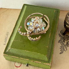Vintage gold tone brooch featuring a cluster of iridescent aurora borealis crystals framed by curved scrolls and rhinestone accents, shown against various backgrounds.