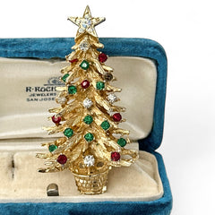 Vintage 1980s gold tone Christmas tree brooch with red, green, and clear rhinestones, featuring a star topper and woven basket base; shown from multiple angles to highlight dimensional detail, condition, and clasp.