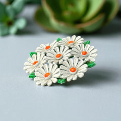Vintage 1960s enamel daisy brooch with white petals, orange centers, and green leaves in a round floral cluster. Silver-toned finish, 2-inch diameter, in very good condition.