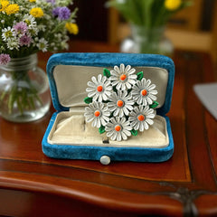 Vintage 1960s enamel daisy brooch with white petals, orange centers, and green leaves in a round floral cluster. Silver-toned finish, 2-inch diameter, in very good condition.