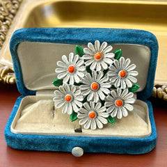 Vintage 1960s enamel daisy brooch with white petals, orange centers, and green leaves in a round floral cluster. Silver-toned finish, 2-inch diameter, in very good condition.