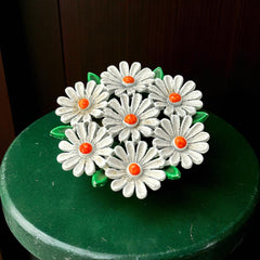 Vintage 1960s enamel daisy brooch with white petals, orange centers, and green leaves in a round floral cluster. Silver-toned finish, 2-inch diameter, in very good condition.