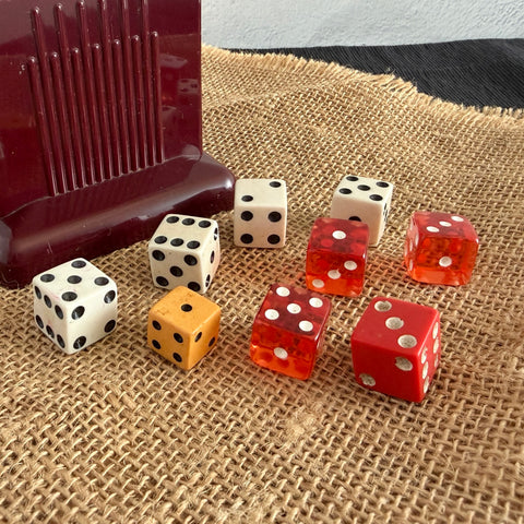 Nine vintage dice in mixed colors and sizes, including a smaller Bakelite die, displayed on textured fabric.