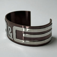 Dolce & Gabbana men's stainless steel cuff bracelet with a brown finish and raised silver-tone logo plaque; modern designer accessory with bold branding and sculptural detail.