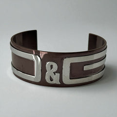 Dolce & Gabbana men's stainless steel cuff bracelet with a brown finish and raised silver-tone logo plaque; modern designer accessory with bold branding and sculptural detail.