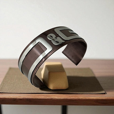 Dolce & Gabbana men's stainless steel cuff bracelet with a brown finish and raised silver-tone logo plaque; modern designer accessory with bold branding and sculptural detail.