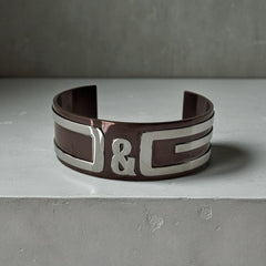 Dolce & Gabbana men's stainless steel cuff bracelet with a brown finish and raised silver-tone logo plaque; modern designer accessory with bold branding and sculptural detail.