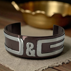 Dolce & Gabbana men's stainless steel cuff bracelet with a brown finish and raised silver-tone logo plaque; modern designer accessory with bold branding and sculptural detail.