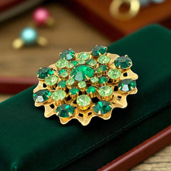 Vintage 1950s gold tone brooch with a ruffled sunburst design, featuring concentric rings of emerald and peridot rhinestones and a large green center stone; bold mid-century costume jewelry with layered sparkle and sculptural detail.