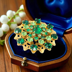 Vintage 1950s gold tone brooch with a ruffled sunburst design, featuring concentric rings of emerald and peridot rhinestones and a large green center stone; bold mid-century costume jewelry with layered sparkle and sculptural detail.