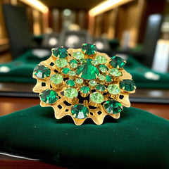Vintage 1950s gold tone brooch with a ruffled sunburst design, featuring concentric rings of emerald and peridot rhinestones and a large green center stone; bold mid-century costume jewelry with layered sparkle and sculptural detail.