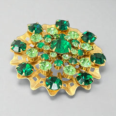 Vintage 1950s gold tone brooch with a ruffled sunburst design, featuring concentric rings of emerald and peridot rhinestones and a large green center stone; bold mid-century costume jewelry with layered sparkle and sculptural detail.